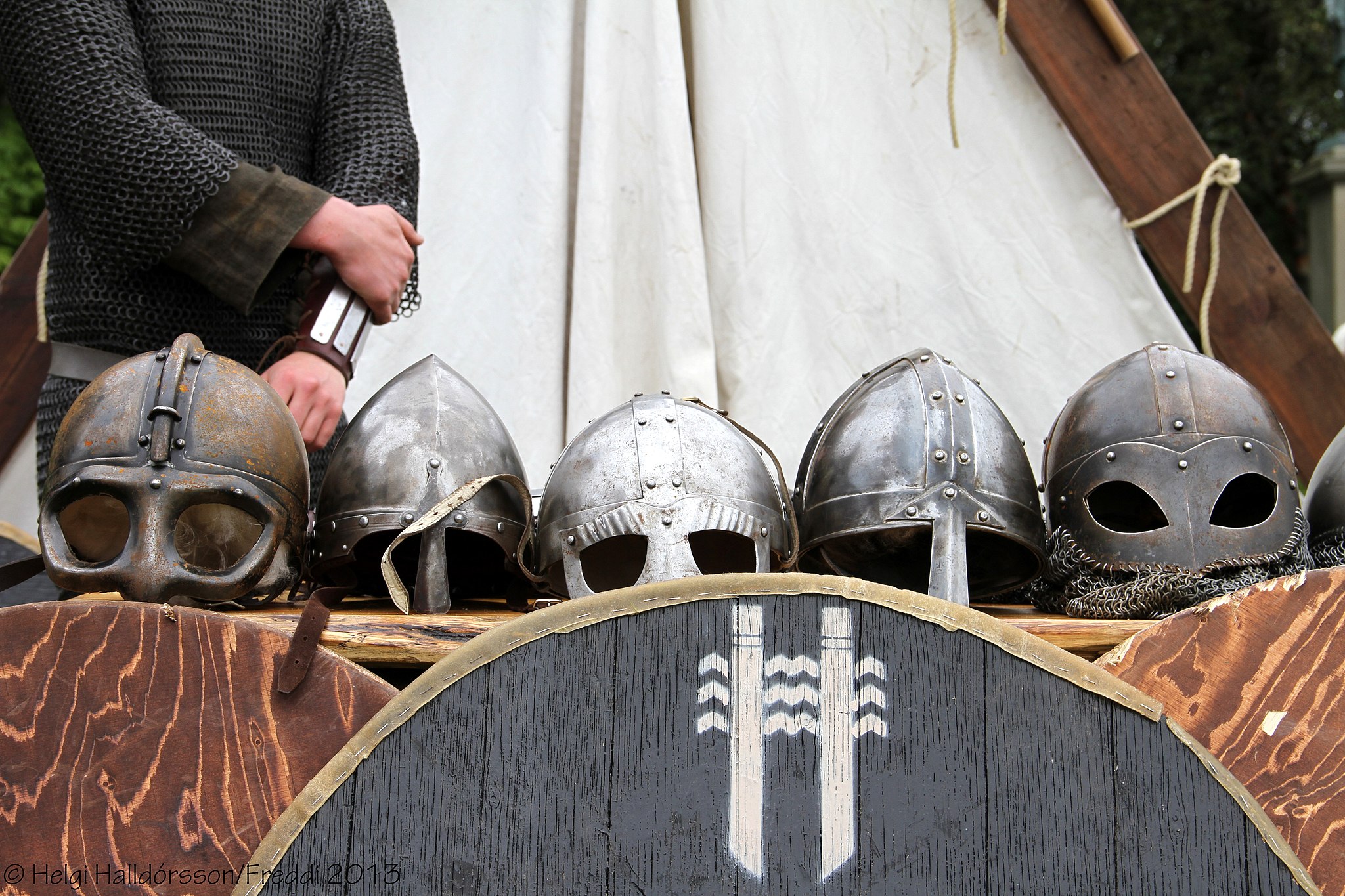 Five Viking helmets sit on a table behind wooden shields