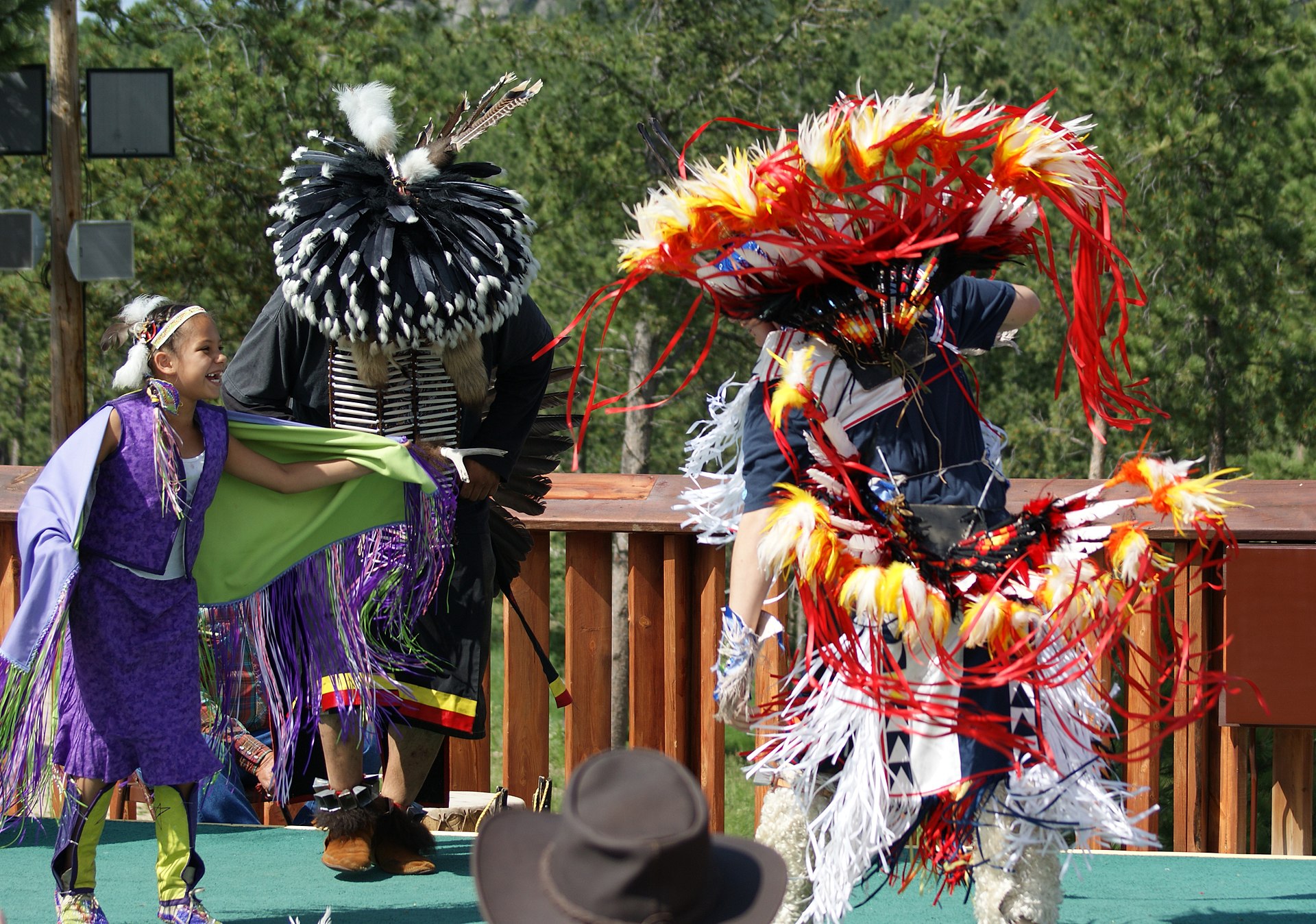 Indigenous dancers perform outdoors; child in purple shawl, men in feathered and ribboned regalia.