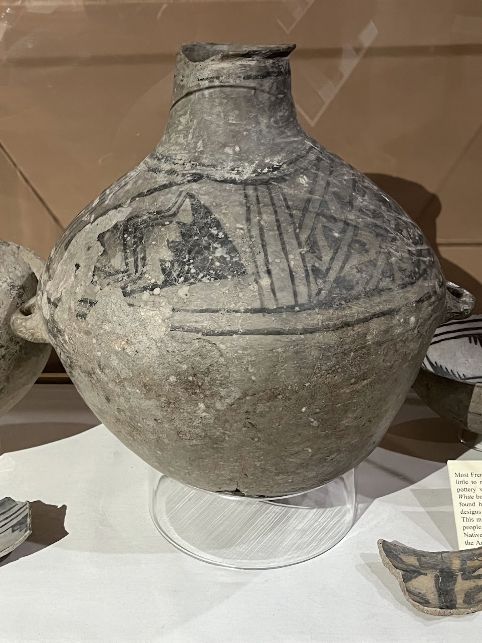 A large, rounded pottery vessel with a narrow neck and small side lugs shows faded triangular and zigzag painted motifs;