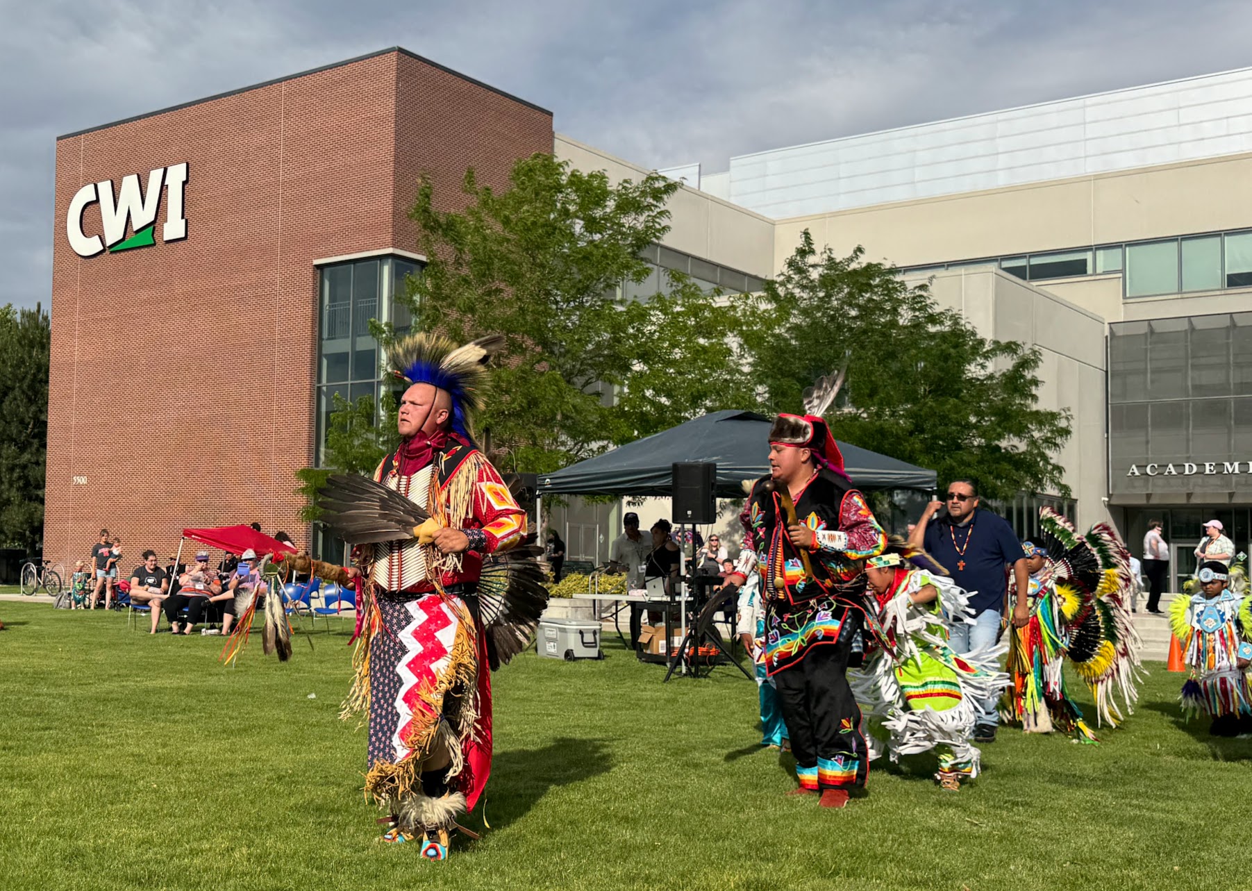 Indigenous dancers in colorful regalia perform outdoors at CWI campus event, with spectators in background.