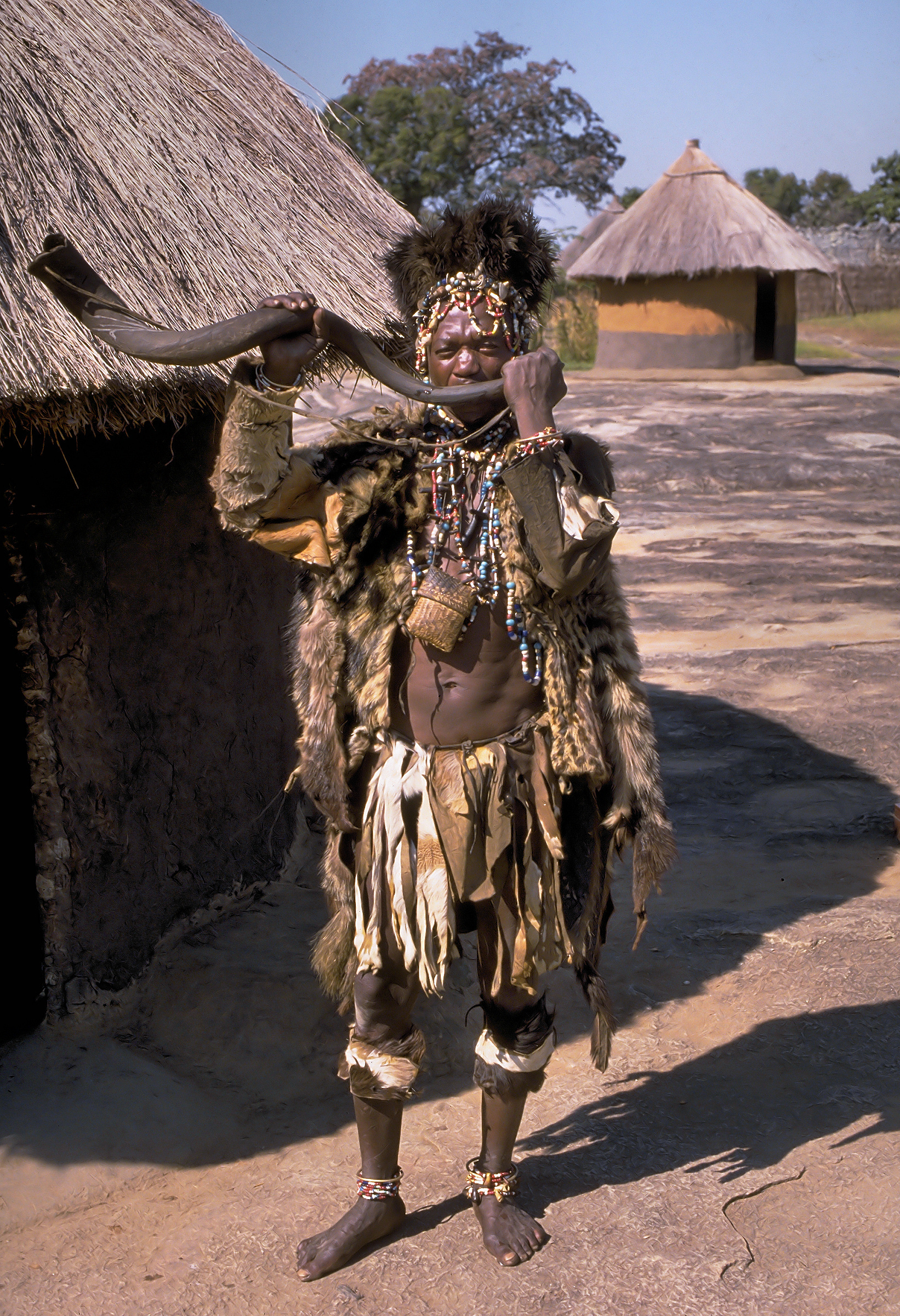a man in ceremonial garb and headdress stands outside a straw hut