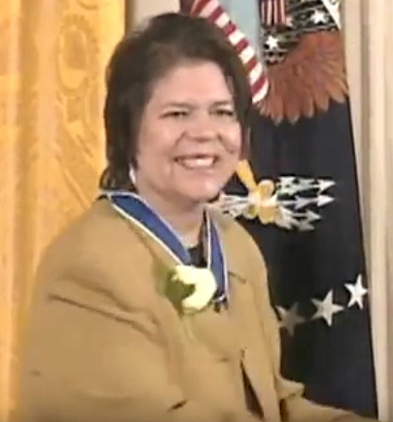 Smiling Native American woman at awards ceremony wearing a medal, U.S. flag and seal behind.