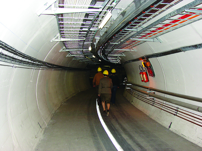 Two photos are shown and labeled “a” and “b.” Photo a shows an aerial view of the Large Hadron Collider. Photo b shows a tunnel of concrete with rails on the ground and tubes and wires running along the wall. Two people walk along the tunnel.