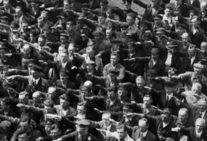 Historical photo of a group of men performing the Nazi salute in Nazi Germany. One person is refusing to do so and standing with his arms crossed.